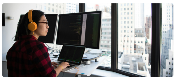 Engineer works on computer code at a standing desk in front of large windows overlooking city